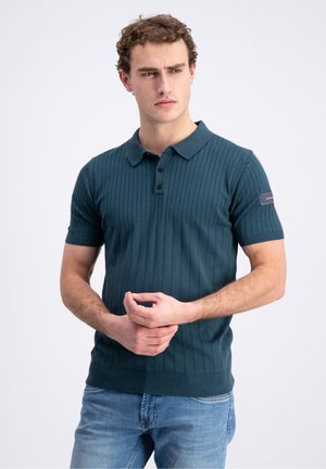 Giovane uomo con capelli ricci che indossa una polo a coste verde petrolio scuro a maniche corte e jeans blu, guarda di lato con le mani congiunte.