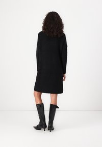 Robe en tricot noire à manches longues et coupe ample, tombant au milieu de la cuisse. Assortie à des bottes à talons en cuir noir à mi-mollet.