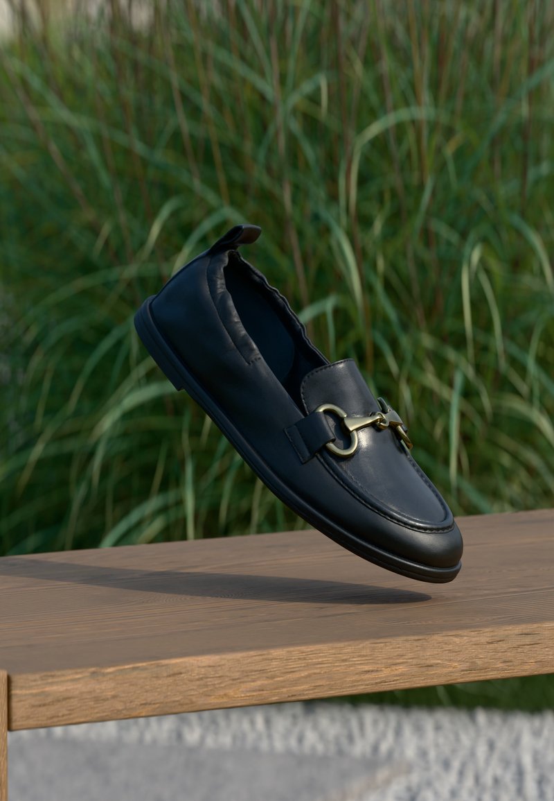 Mocassin en cuir noir avec détail en métal doré flottant au-dessus d'un banc en bois, fond d'herbe verte.
