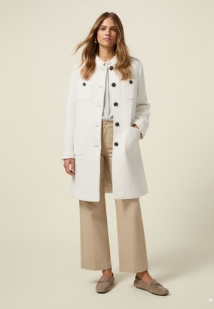 Witte textuur jas met een knopen-front, vier zakken en zwarte knopen, gecombineerd met beige brede broek en lichtgrijze mocassins.