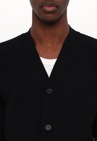 Cardigan noir avec un décolleté en V, tissu texturé et trois boutons noirs à l'avant, superposé sur une chemise blanche.