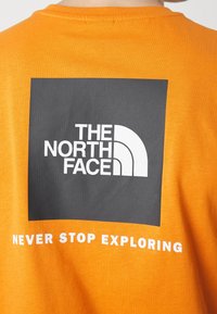Oranžové bavlněné tričko s velkým černým obdélníkovým grafickým prvkem na zadní straně s bílým textem a logem "THE NORTH FACE" a "NEVER STOP EXPLORING."
