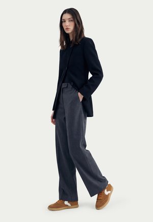 Woman wearing a black blazer, dark gray wide-leg pants, and brown sneakers, standing with one hand in pocket against a plain background.