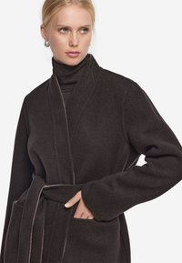 Mörkbrun ullkappa med bälte, vida ärmar, sjalkrage och sidofickor. Har en mjuk textur och subtil kontrastsöm.