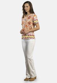 Blusa de manga corta con patrón colorido y escote en V, que presenta diseños florales y geométricos, combinada con pantalones blancos y botas de tacón en color beige.