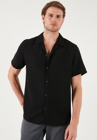 Camisa negra de manga corta con cuello, cierre de botones en la parte delantera y una textura suave. Presenta un corte relajado y un diseño minimalista.