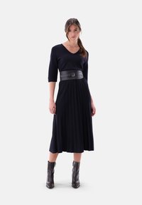 Marineblaues plissee Kleid mit V-Ausschnitt und Dreiviertelärmeln, tailliert mit einem schwarzen Ledergürtel. Das Model trägt schwarze Knöchelstiefel.