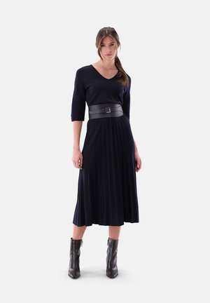 Marineblaues plissee Kleid mit V-Ausschnitt und Dreiviertelärmeln, tailliert mit einem schwarzen Ledergürtel. Das Model trägt schwarze Knöchelstiefel.