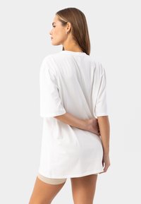Camiseta blanca de gran tamaño con mangas cortas, hecha de una tela suave. Detalle de costura en la parte trasera notable. Combina con pantalones cortos beige.