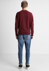 Vínová mikina s dlouhými rukávy, žebrovanými manžetami a lemem, v kombinaci s světle modrými džínami a černými teniskami s bílými akcenty.