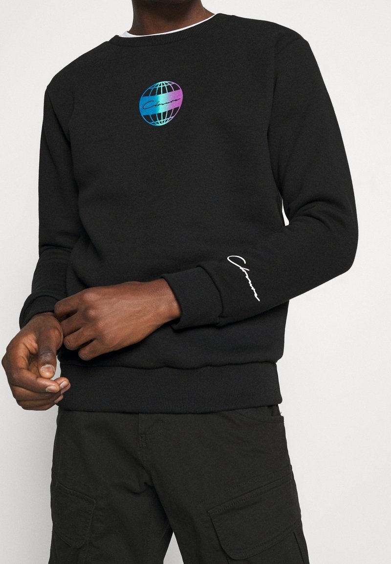 Homme portant un sweat-shirt noir avec un logo globe bleu et violet sur la poitrine et un logo script blanc sur la manche gauche, associé à un pantalon cargo noir.