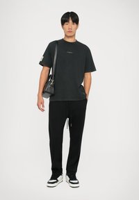 Sort T-shirt med et grønt logo, afslappet pasform; parret med sorte bukser med snoretræk og sorte sneakers. En lille sort crossbody-taske bæres.