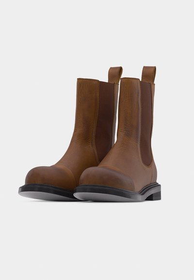 Bottes basses en cuir marron avec panneaux latéraux élastiques, languettes de tirage, bouts arrondis et semelles noires, présentées sur un fond clair.