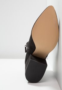 Bottine noire en daim avec semelle beige pointue, talon bloc et fermeture éclair latérale, présentée vue de la semelle sur fond blanc.