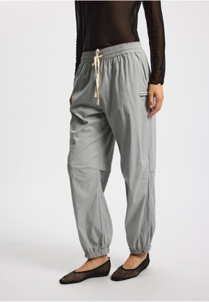 Pantalones gris claro, holgados, con cintura elástica, cordón ajustable y puños elásticos. Cuentan con un bolsillo lateral y una pequeña etiqueta en la parte frontal.