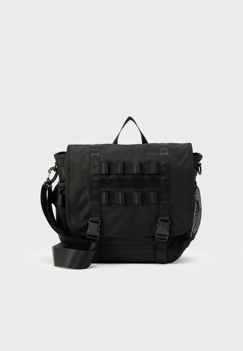 Μαύρη ταξιδιωτική τσάντα τακτικής χρήσης με ρυθμιζόμενο ιμάντα ώμου, εμπρόσθιες αγκράφες, θηλιές και δίχτυνη πλευρική τσέπη σε λευκό φόντο.