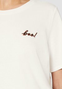 T-shirt en coton blanc avec un col arrondi et des manches courtes, orné d'un texte brodé noir et orange "boo!" sur la poitrine gauche.