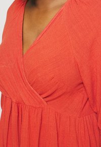 Robe rouge texturée avec un décolleté en V, une taille froncée et une jupe fluide. Le tissu présente un motif de tissage subtil avec un drapé doux.