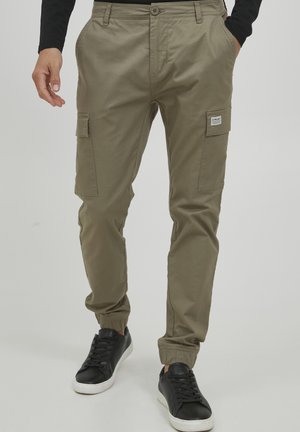 Homme portant un pantalon de jogging cargo kaki avec des poignets élastiques et des baskets noires, les mains dans les poches, debout sur un fond gris.