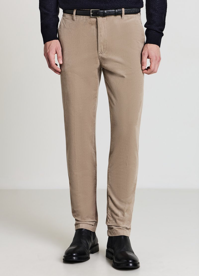 Man wearing beige slim-fit trousers with black belt, black sweater, and black leather ankle boots, standing on plain background.