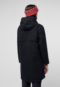 Cappotto impermeabile nero con cappuccio, dotato di una fessura sul retro e un orlo leggermente più lungo. Il modello indossa una fasciatura per capelli in maglia multicolore.