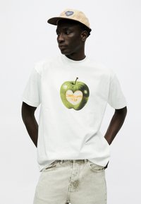 Άνδρας με μπεζ καπέλο, λευκό μπλουζάκι με πράσινο μήλο και ξεβαμμένο μπεζ τζιν, στέκεται με τα χέρια στις τσέπες μπροστά σε λευκό φόντο.