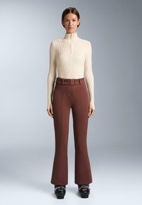 Krém színű bordázott garbós pulóver, cipzárral, barna trapéz nadrággal párosítva, amely övvel ellátott magas derekú és kontrasztos varrási részletekkel rendelkezik.