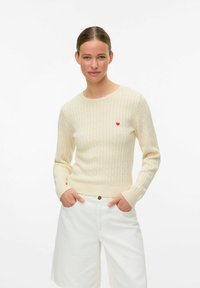 Lichtbeige geribde trui met lange mouwen, voorzien van een klein rood hartembleem op de linkerborst. Gepaard met witte shorts.