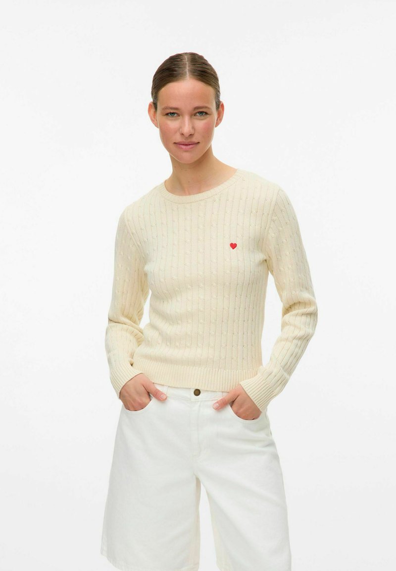 Lichtbeige geribde trui met lange mouwen, voorzien van een klein rood hartembleem op de linkerborst. Gepaard met witte shorts.