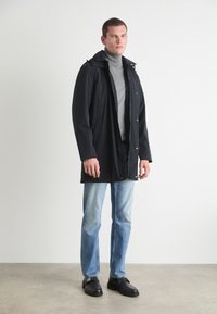 Melnā ūdensnecaurlaidīgā jaka ar kapuci, pogu aizdari un divām sānu kabatām, uzvilkta virs gaiši pelēkas turtleneck vestes un zila džinsu biksēm. Melnas kurpes.