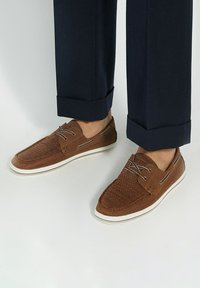 Brown woven casual shoes with a textured upper, white rubber sole, and decorative laces. Paired with dark cuffed trousers.