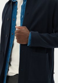 Námořnický cardigan s texturovaným pletením, pod ním modrá denimová košile, detaily s knoflíky a dvěma předními kapsami. Minimalistický design.