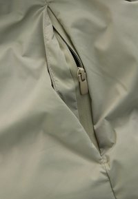 Bežová tkanina so zipsovým vreckom s jemným leskom. Zips je svetlo béžový, čím dopĺňa hladkú textúru materiálu.