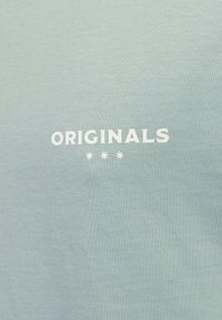 Svetlomodrá bavlnená tkanina obsahuje slovo "ORIGINALS" v tučnom bielom texte s štyrmi malými hviezdičkami pod ním. Hladká, rovnomerná textúra.