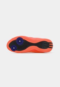 Helfärgade orange sportskosulor med svart texturerad greppyta och två runda blå greppdynor nära hälen mot en vit bakgrund.