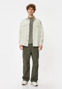Lysbeige skjorte med knapper over en grå t-shirt, parret med olivengrønne cargo-bukser. Skoene er hvide med brune detaljer.
