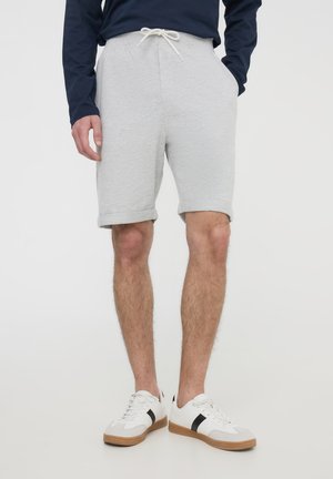Uomo che indossa pantaloncini grigio chiaro con orlo arrotolato, camicia a maniche lunghe blu navy e sneakers bianche con striscia nera, in piedi con le mani nelle tasche.