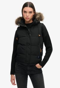 Schwarze, gefütterte Weste mit einer Kapuze aus Fell, mit einem Reißverschluss vorne, braunen Toggle-Details und einem gesteppte Design in einer taillierten Form.