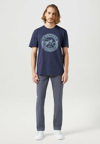 Marineblauwe katoenen t-shirt met een rond logo met een motorontwerp, gecombineerd met grijze slim-fit broek en witte sneakers.