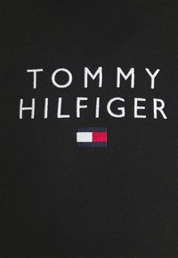 Tissu noir avec le texte "TOMMY HILFIGER" brodé en blanc et un petit logo rectangulaire en marine, blanc et rouge sous le texte.