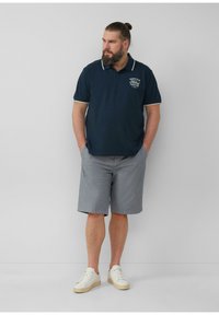 Navyblaues Poloshirt mit weißem Kragen und Logodetail, kombiniert mit grauen karierten Shorts und weißen Sneakers, die ein lässiges Outfit präsentieren.