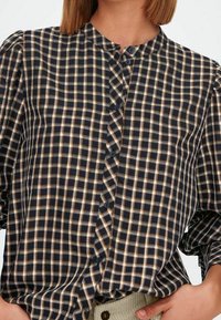 Camicia a quadri blu navy con check crema e marrone, colletto a fascia e maniche a sbuffo, con una chiusura a bottoni verticale.