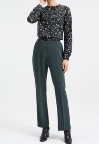 Halenka s květinovým vzorem a dlouhým rukávem v námořnické modré barvě s mnoha barevnými akcenty, spárovaná s tmavě zelenými kalhotami s vysokým pasem a černými kotníkovými botami na podpatku.