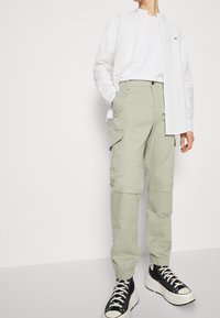 Ljusgröna cargobyxor med elastiska muddar, sidofickor och en jämn yta, tillsammans med en vit skjorta med knappar och svarta sneakers.