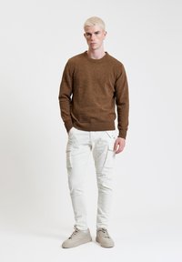 Brązowy sweter o fakturze, białe spodnie typu cargo z kieszeniami i beżowe sneakersy. Model stoi na tle jednolitego jasnego tła.