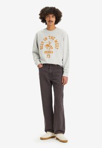 Sweatshirt gris clair avec un texte graphique orange, associé à un jean large gris foncé et des baskets blanches avec des accents marron.