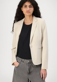 Beige bleiseri, jolla on slim fit -muotoilu, yksisoljinen kiinnitys ja kärjelliset läpät, yhdistetty mustaan paitaan ja harmaisiin korkeavyötäröisiin housuihin.