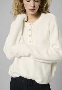 Weißer Strickpullover mit Rundhalsausschnitt, langen Ärmeln und fünf dekorativen Knöpfen an der Vorderseite. Weiche Textur und lockere Passform.