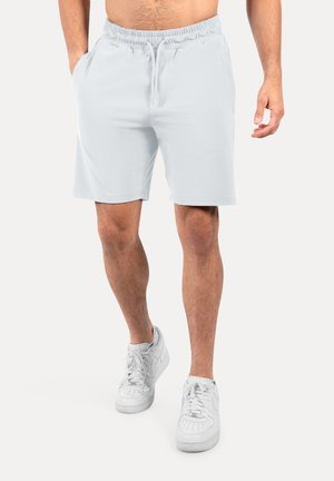 Hombre con pantalones cortos blancos lisos con cordón y zapatillas blancas, de pie con una mano en el bolsillo contra un fondo blanco.