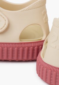 Plastic sandals in cream and pink, featuring a ridged sole, circular fastening, and side cutouts for ventilation. Textured upper for grip.
